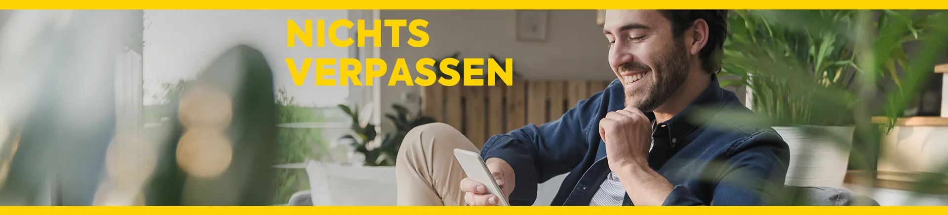 Junger Mann sitzt am Sofa und liest lächelnd auf seinem Smartphone. Text "Nichts verpassen" in gelber Schrift auf dem Foto.
